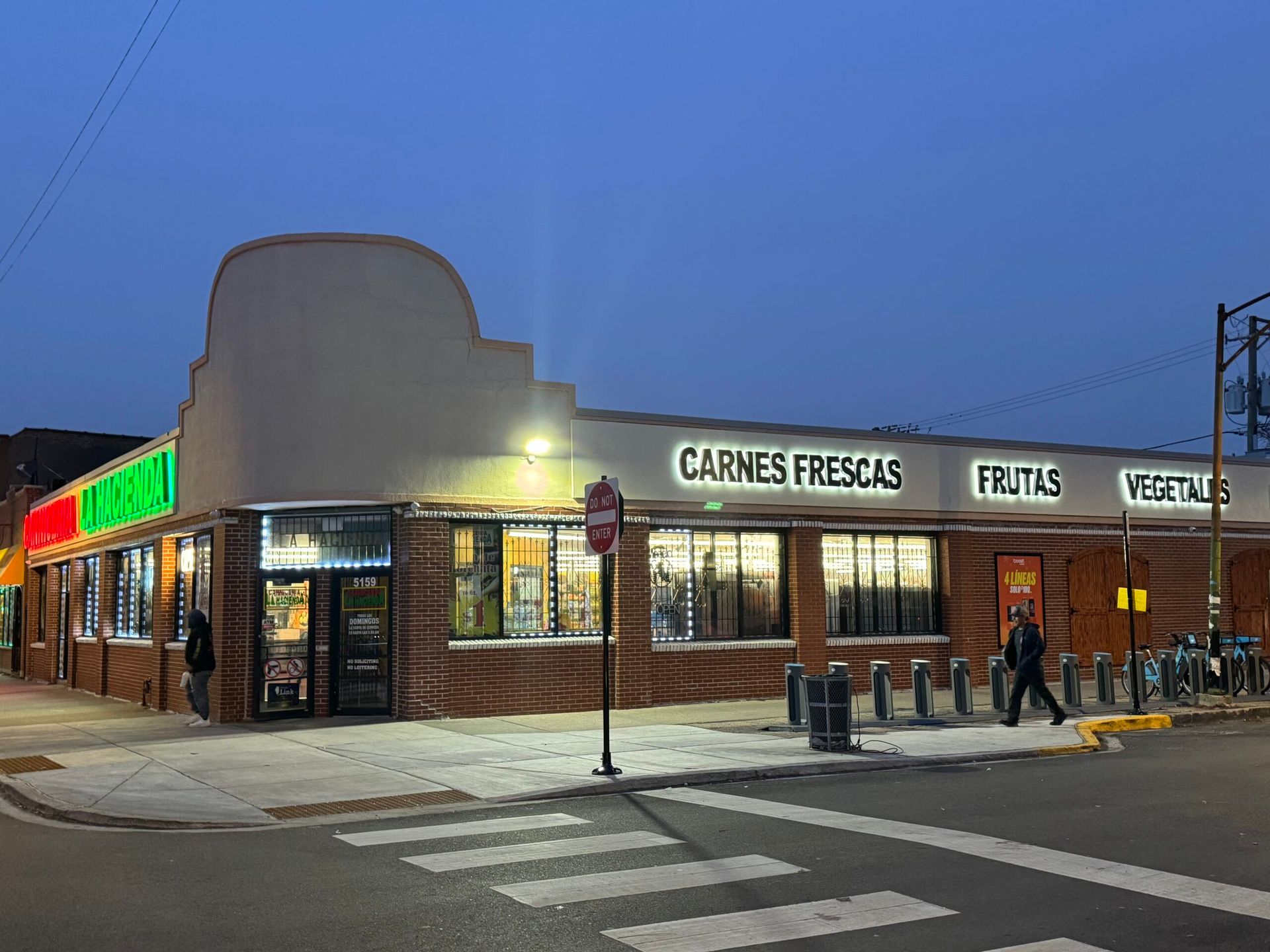 Corner grocery store at dusk, lit storefront with Spanish signs for meats, produce, and tortillas.