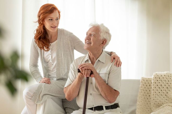 Woman helping an elderly man with a cane, both smiling on a bright couch.