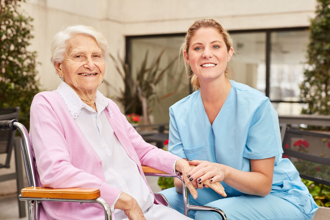 Caregiver in blue scrubs smiling beside an elderly woman in a wheelchair outdoors.