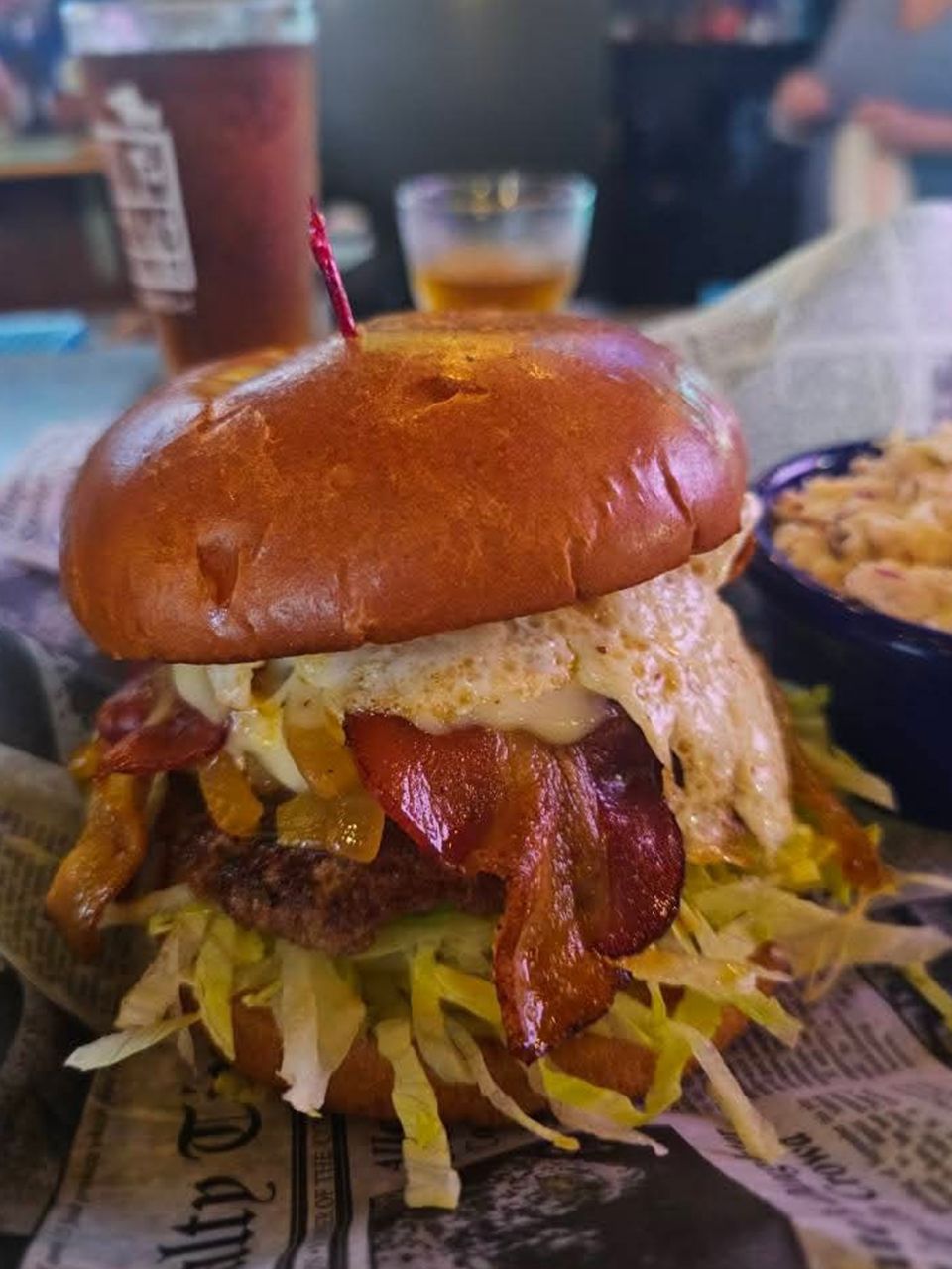 Burger with bacon, fried egg, and lettuce on a brioche bun, with a side of mac and cheese and a beer.