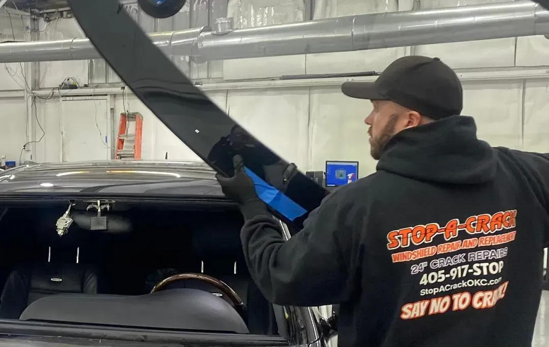 Man installing auto part on a black car in a shop, wearing black hat, gloves, and sweatshirt with 