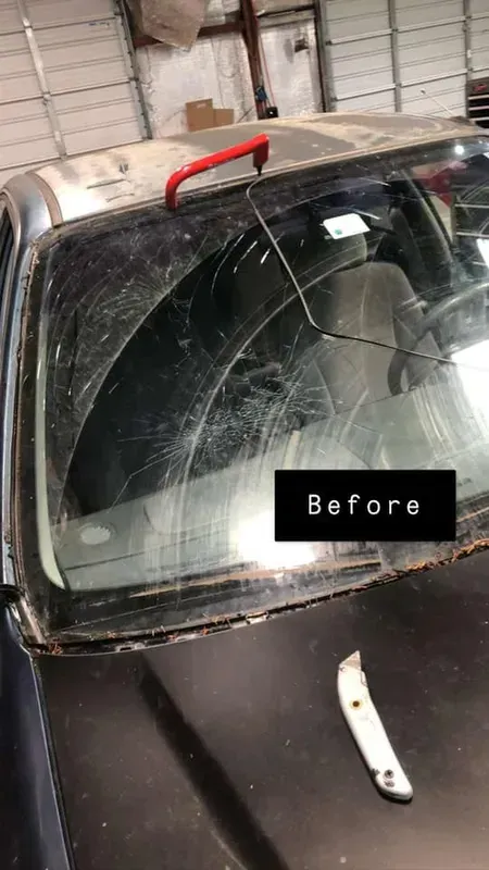A car windshield heavily damaged with cracks. A red handle tool rests on top, a utility knife below. 