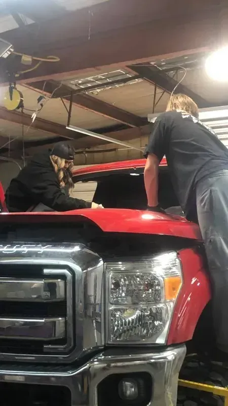 Two people working on the hood of a red truck in a garage.