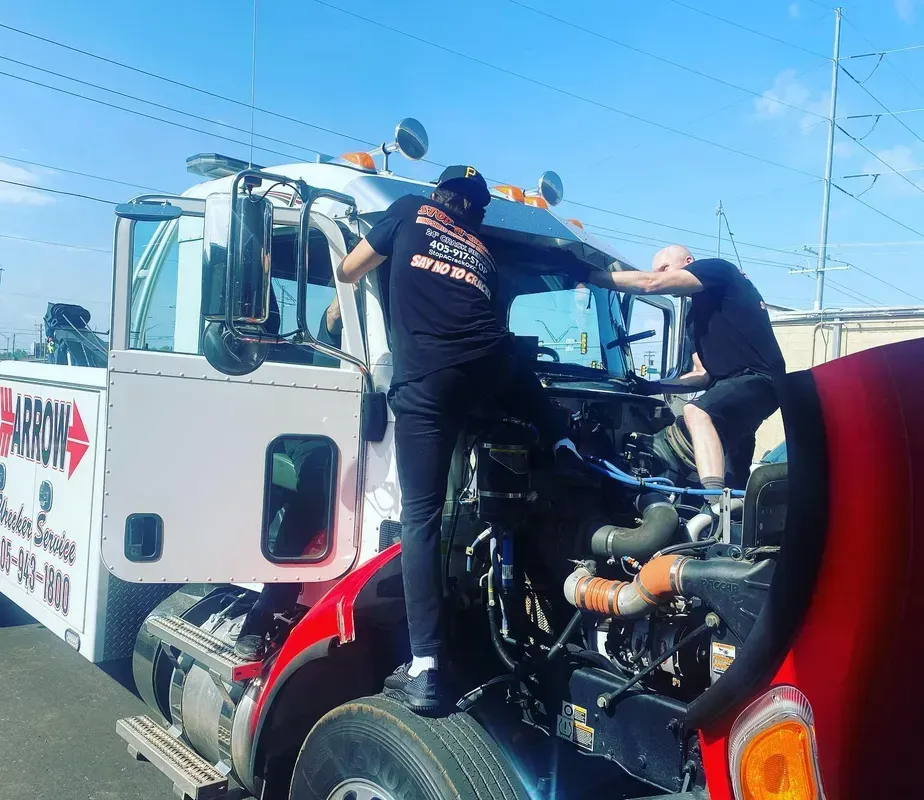 Two people working on a tow truck's engine. One stands on the bumper, the other leans in. Bright sunlight.