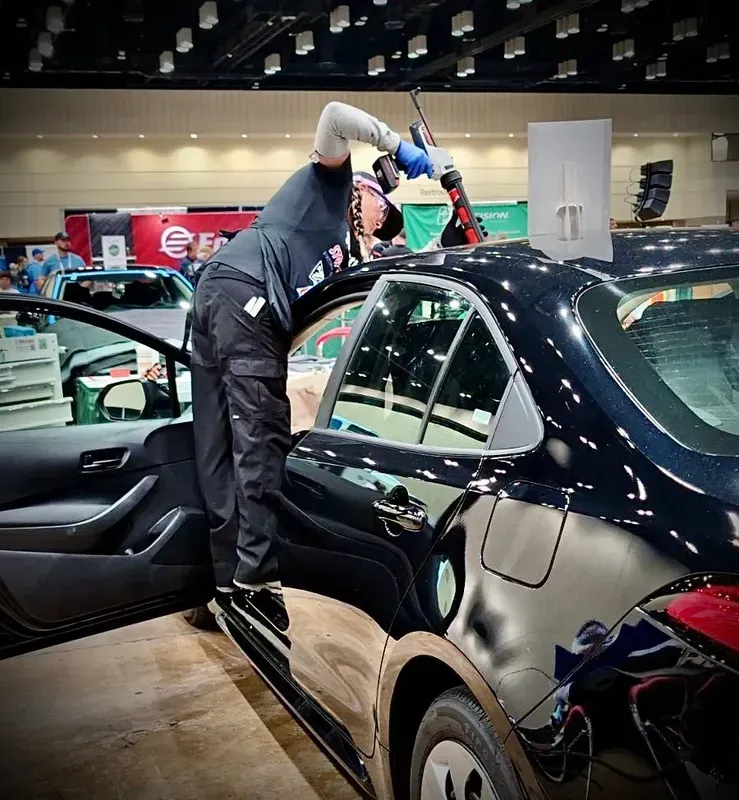 Man using tool on black car, standing on running board in a convention hall.