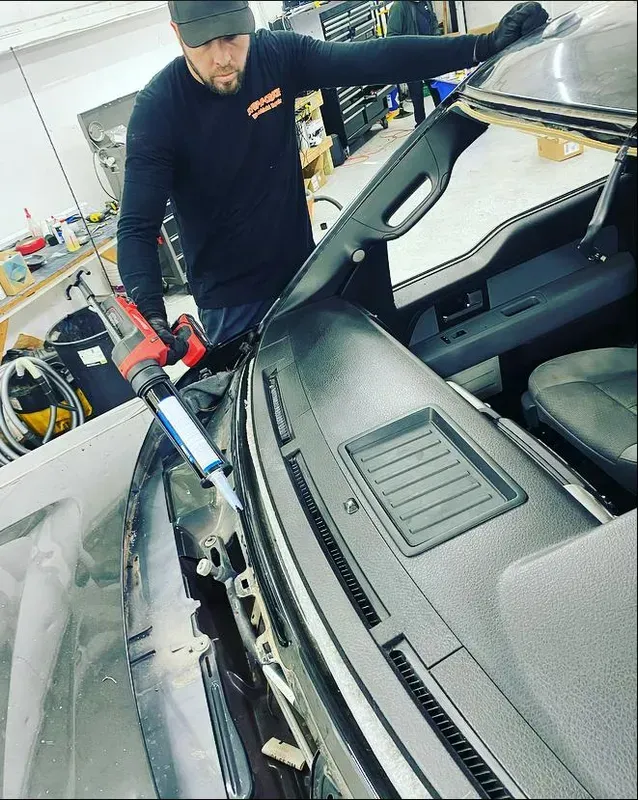 Man using a caulking gun to apply adhesive to a car windshield frame. Inside a shop.