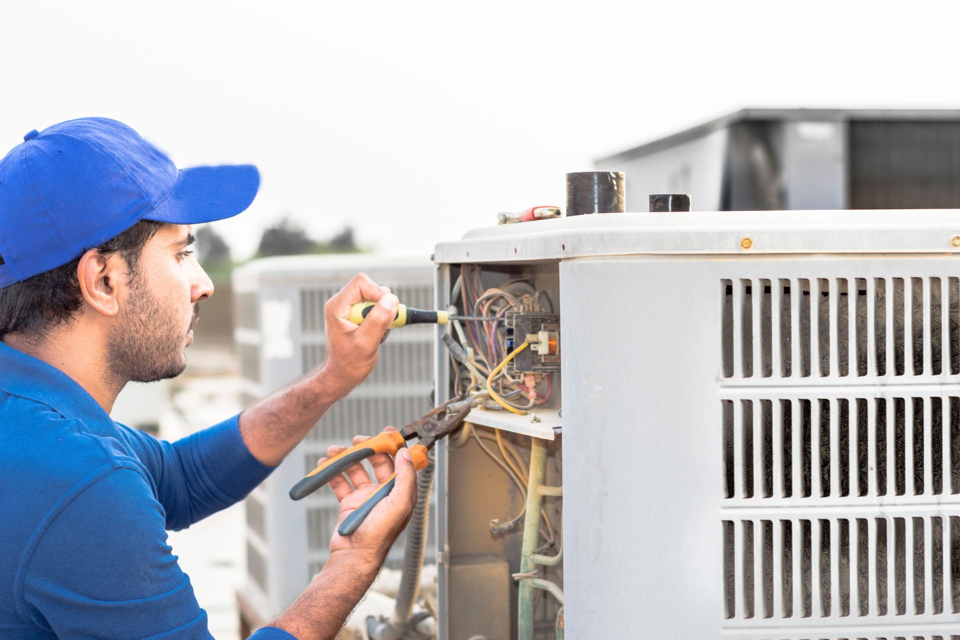 HVAC technician repairs an outdoor AC unit with pliers and a screwdriver on a rooftop.