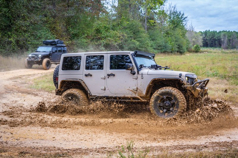 A white jeep is driving through the mud on a dirt road.