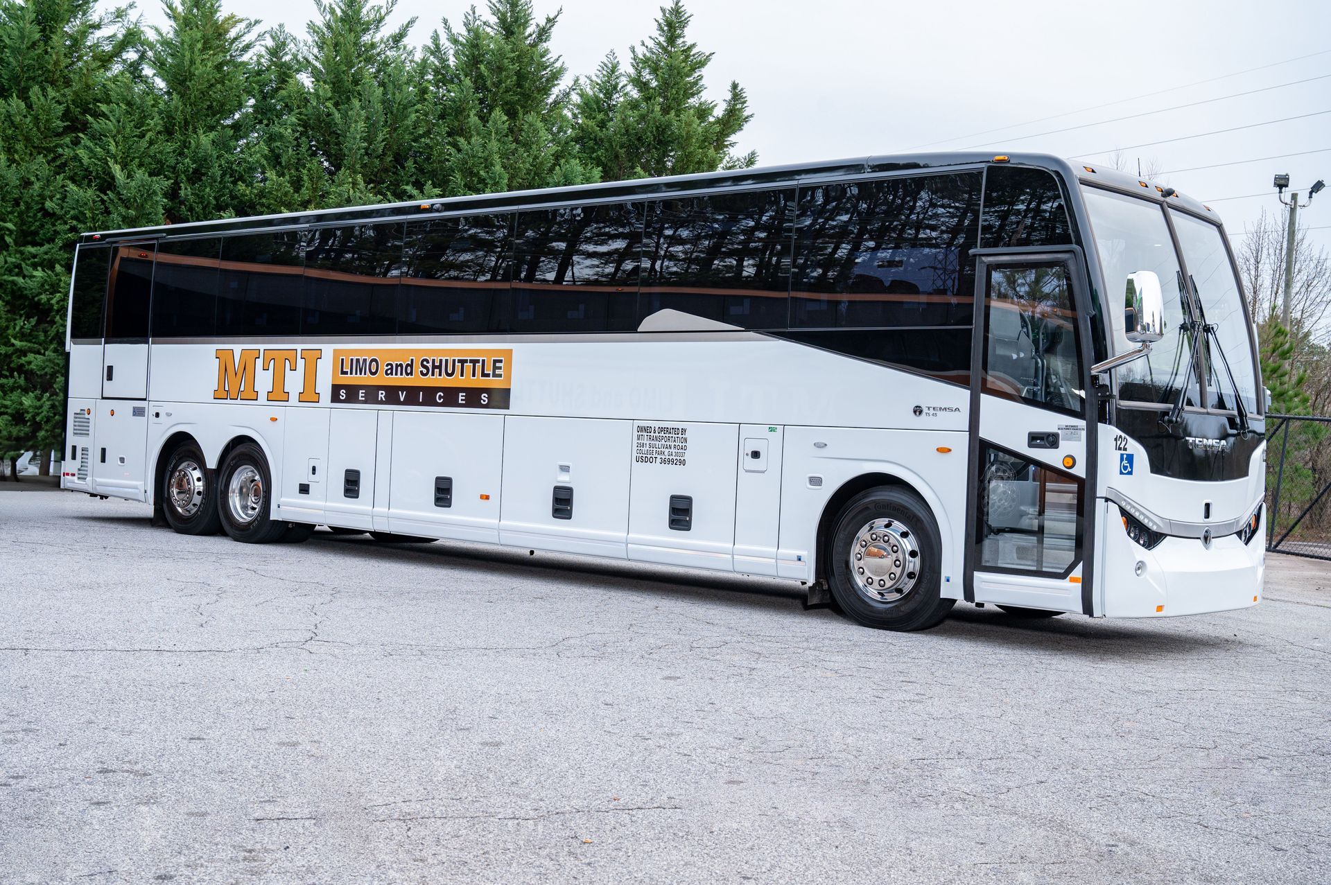 White luxury coach bus with tinted windows parked on a gravel lot beside trees