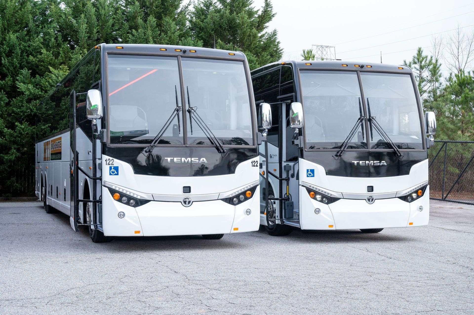 Two white coach buses parked side by side on a gravel lot with trees in the background