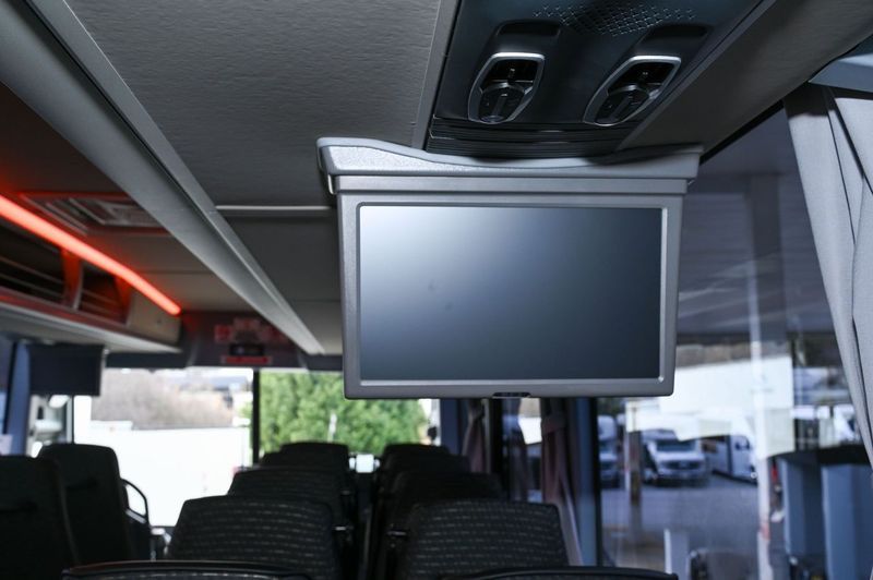 Ceiling-mounted monitor inside a bus or shuttle, with rows of seats below.
