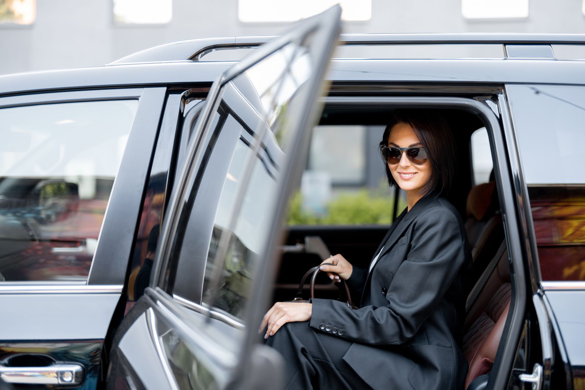 Woman in sunglasses sitting in the back seat of a black car, door open.