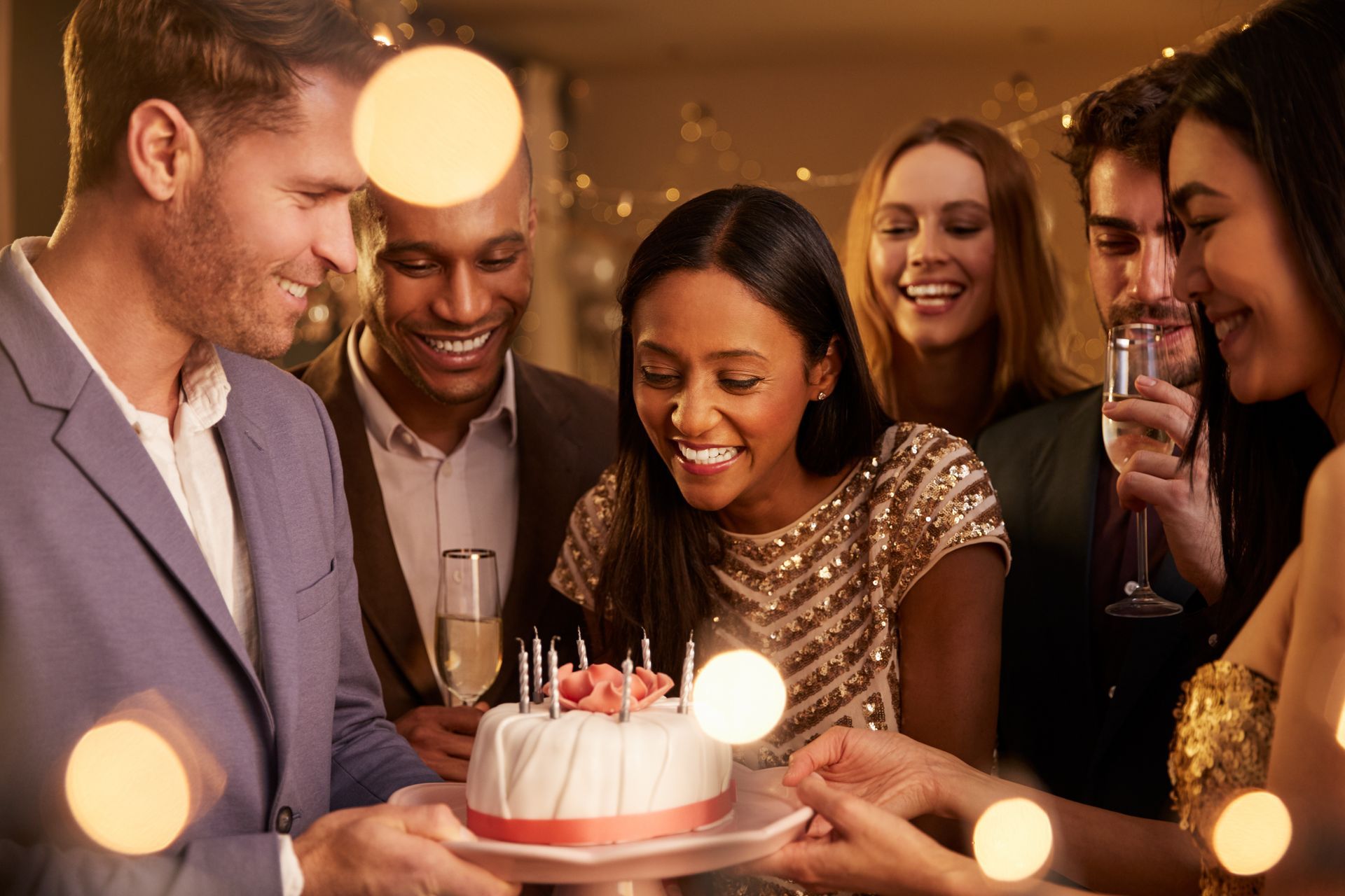 Friends gathered around a birthday cake, smiling and celebrating in a warmly lit room.