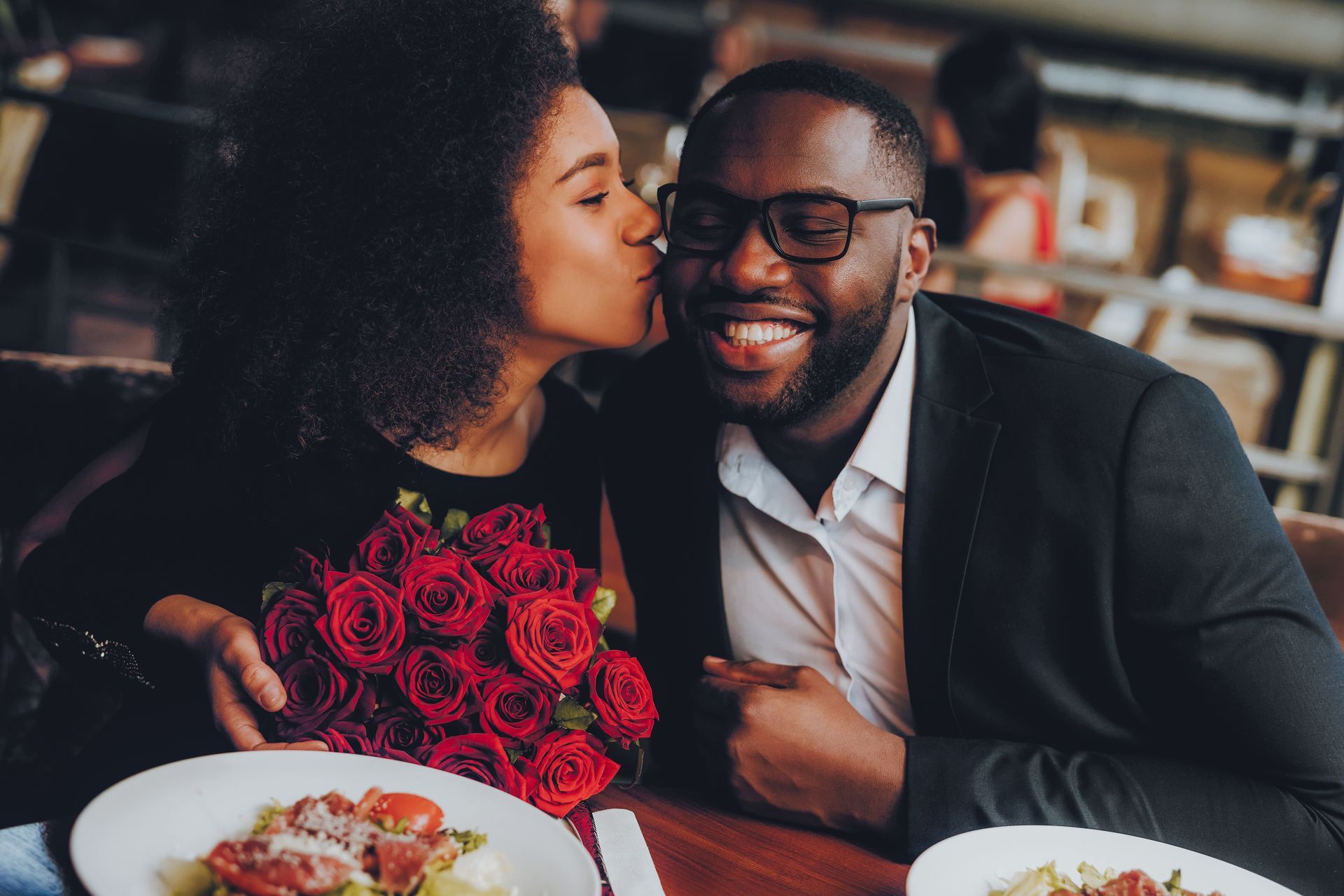 Couple sharing a kiss at a restaurant table with a bouquet of red roses