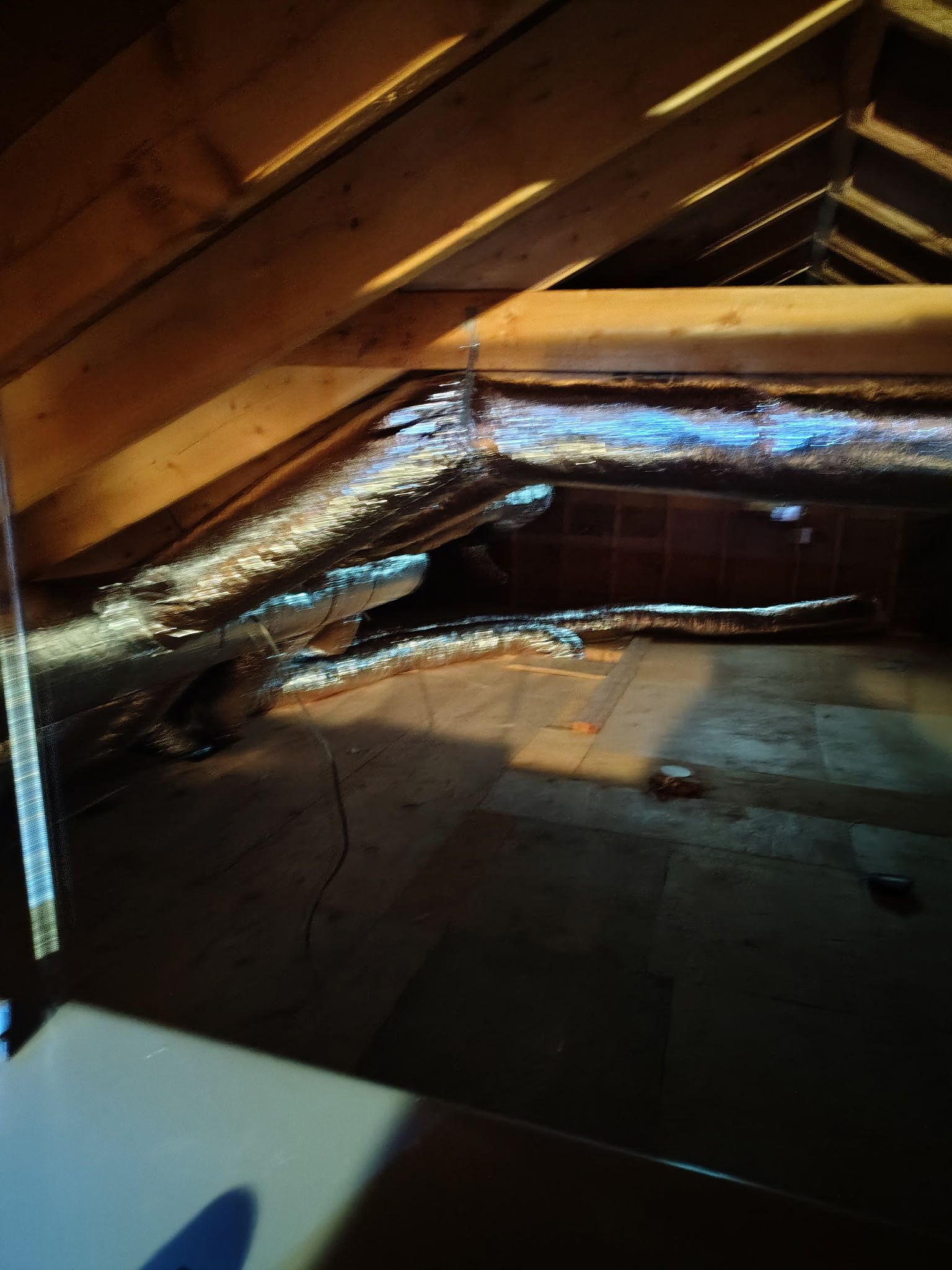 Inside an attic: silver ductwork snaking between wooden rafters and a dark, unfinished floor.