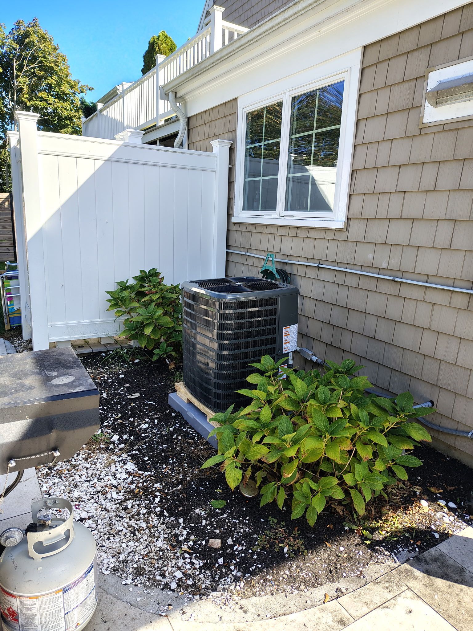 Air conditioning unit next to a house with white fence, landscaping, and window.