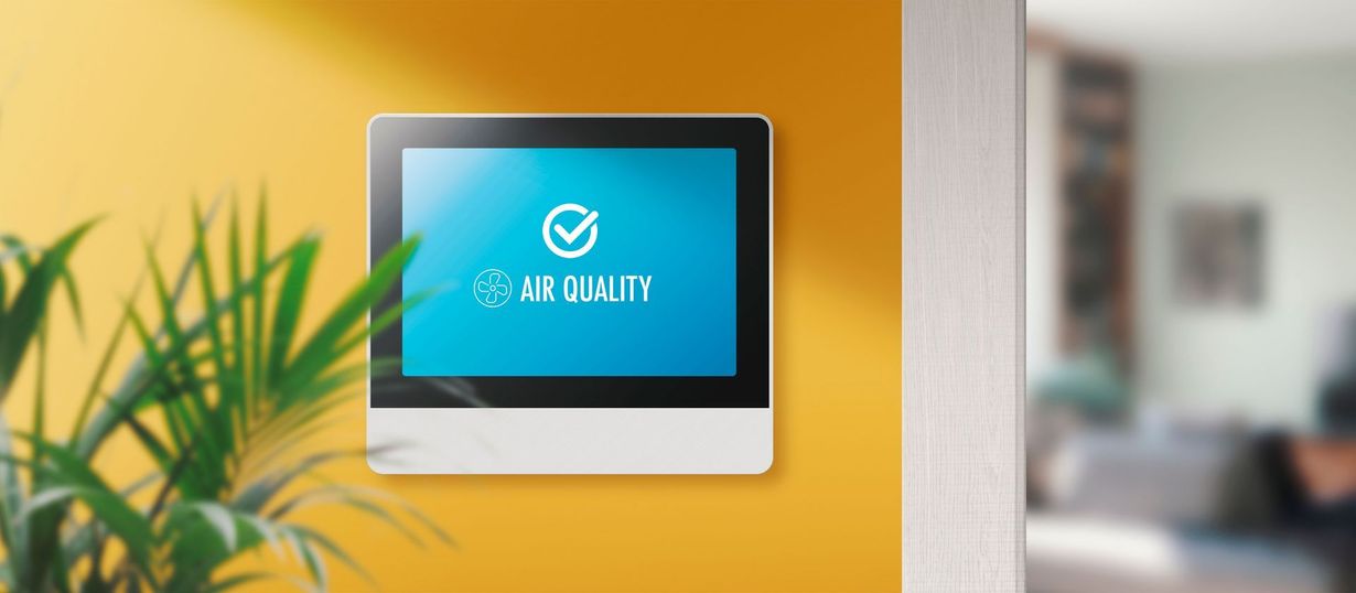 Air quality monitor on a yellow wall with text "Air Quality" displayed. A plant is in the left foreground.