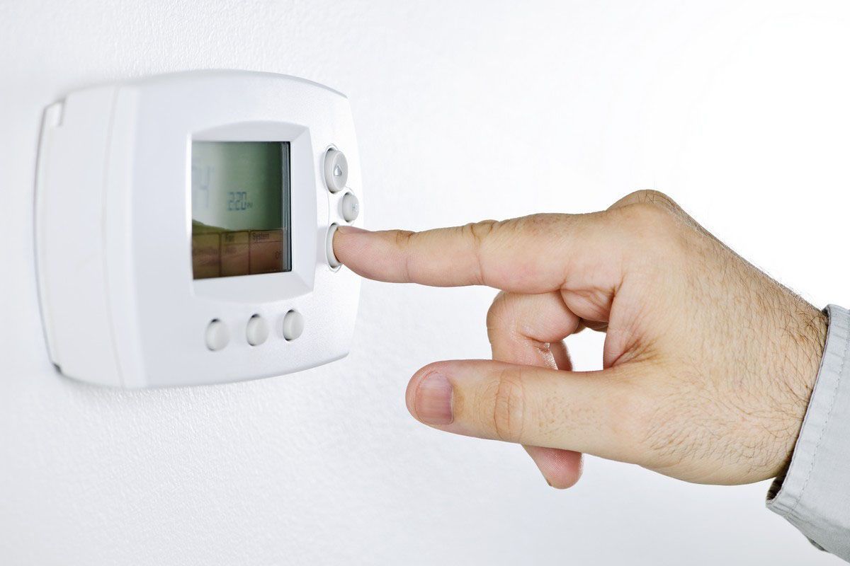 Hand pressing a button on a white thermostat mounted on a wall.
