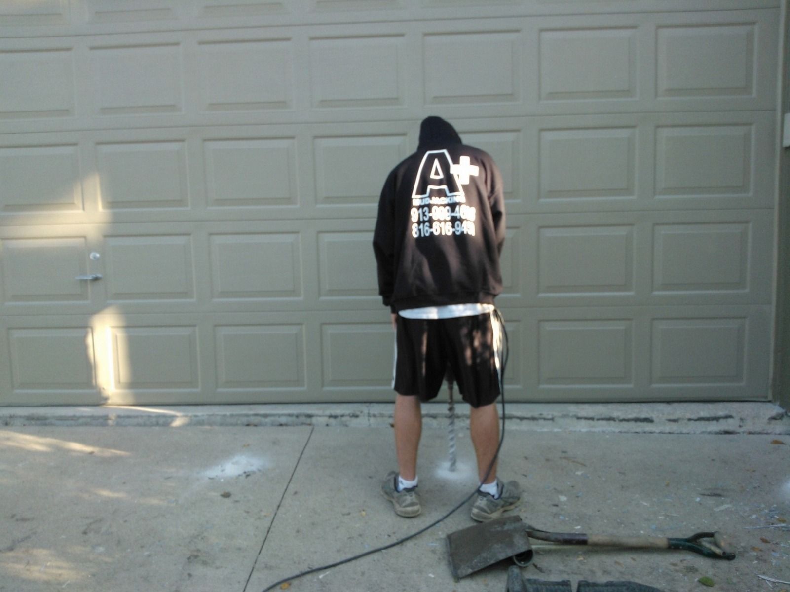 A man is standing in front of a garage door wearing a black hoodie with the letter a on the back