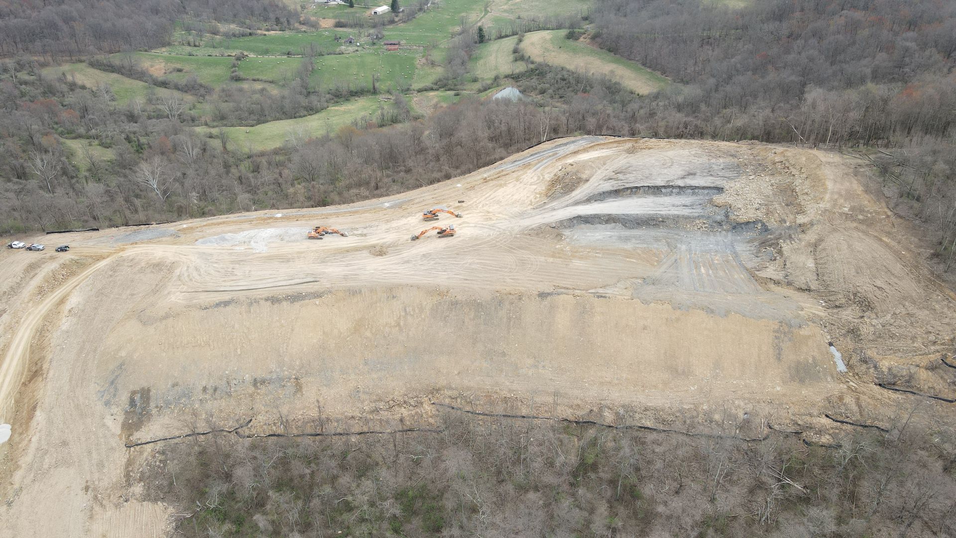 Aerial view of a barren, terraced hillside with exposed soil beside green fields and forested slopes