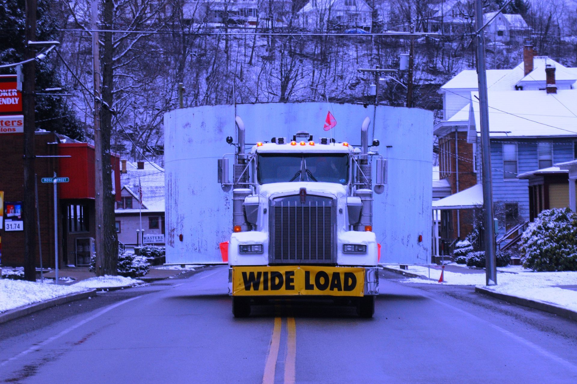 White truck with “WIDE LOAD” sign blocking a snowy street between houses and storefronts