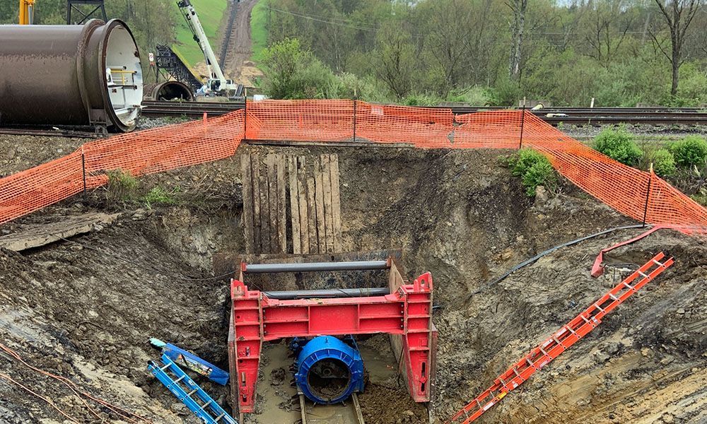 Construction trench with red shoring, blue pipe, orange safety fencing, and heavy equipment above ground