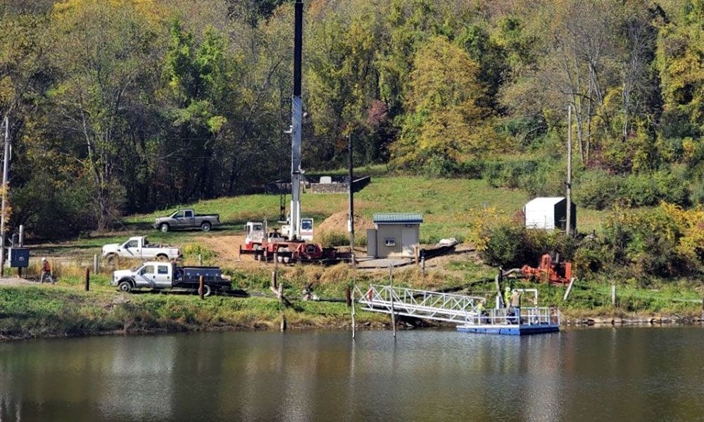 Lakeside worksite with trucks, equipment, and a small dock beside trees in autumn