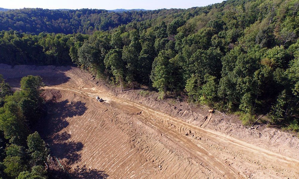 Aerial view of a dry, cleared dirt road cutting through dense green forest on a hillside.