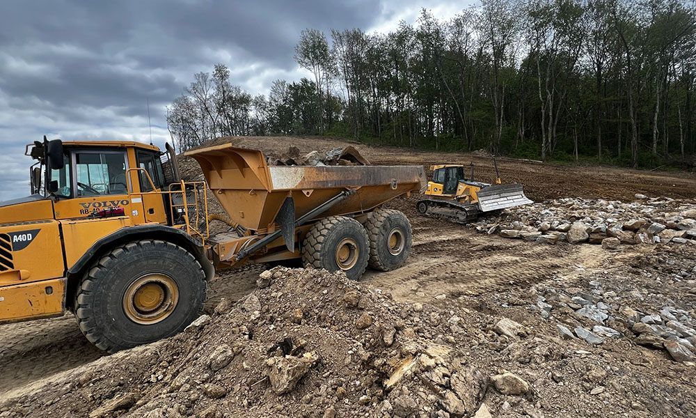 Yellow dump truck hauling dirt at a rocky construction site with trees in the background