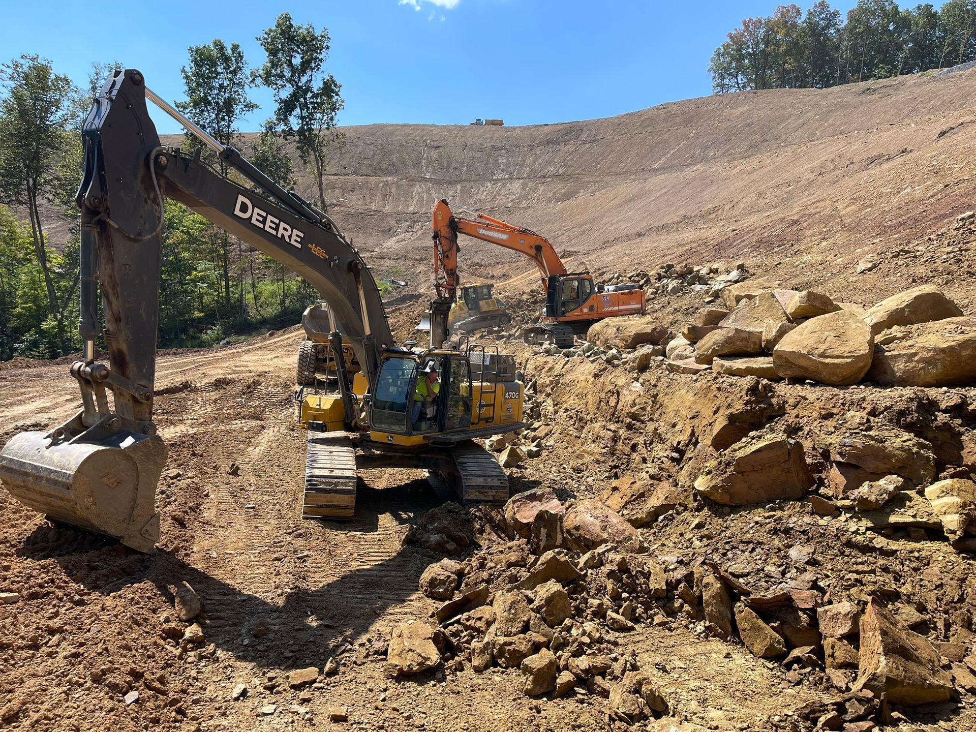 Excavator loading rocks at a dry hillside construction site under a blue sky