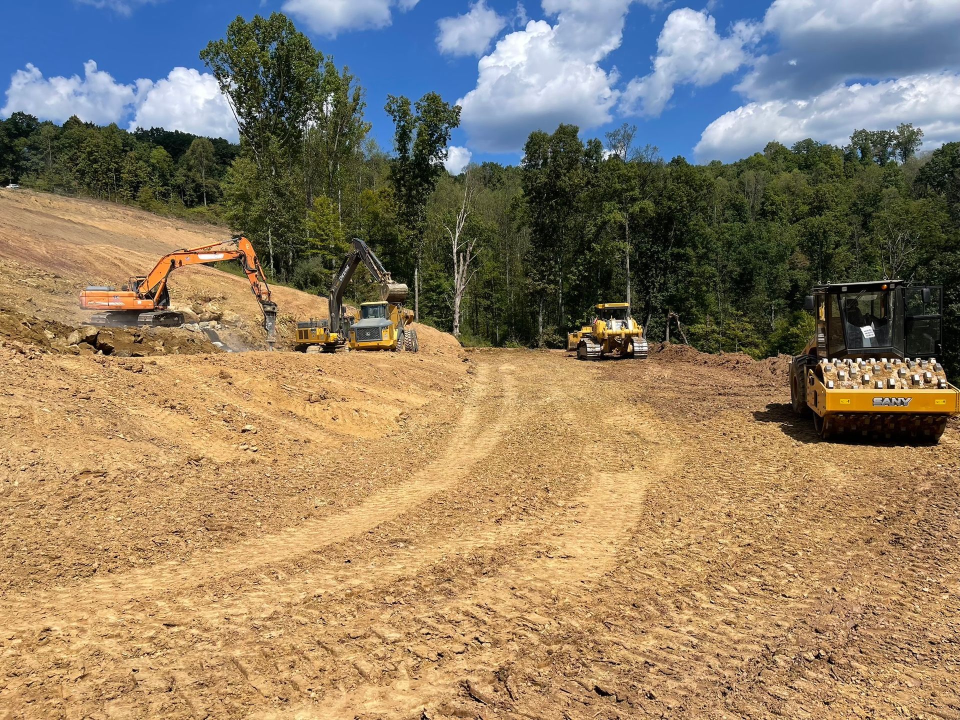 Construction site with excavators and a bulldozer on a dirt clearing beside trees under a blue sky