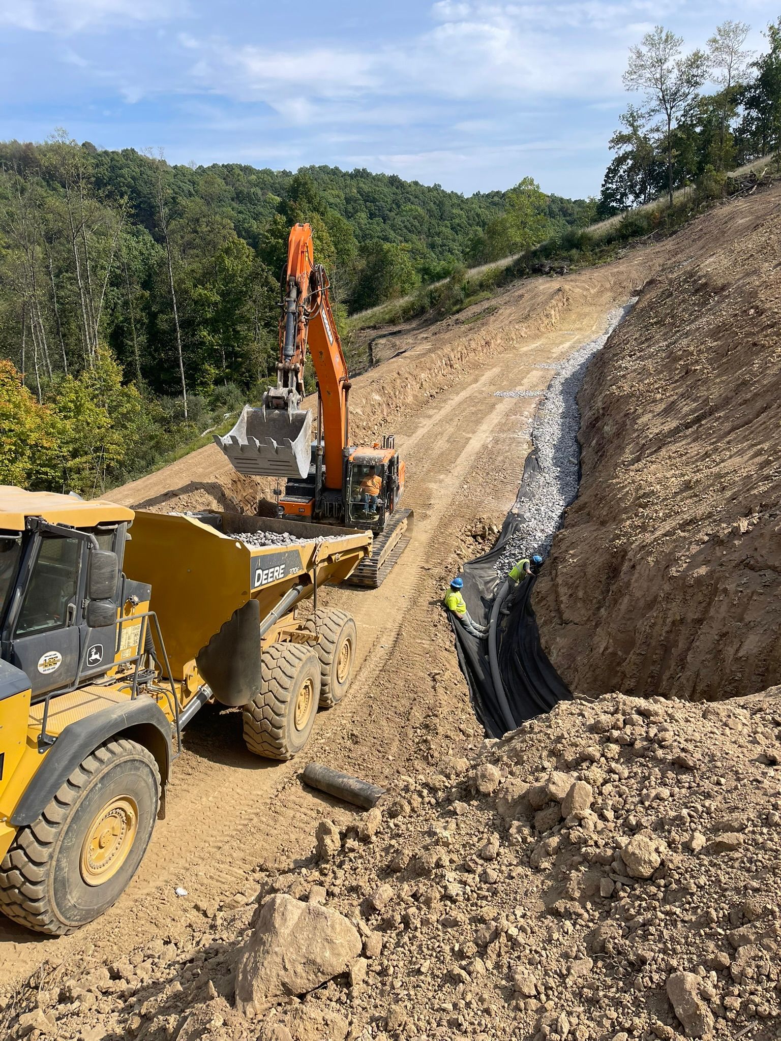 Construction vehicles excavating a dirt hillside beside a steep road and large trench