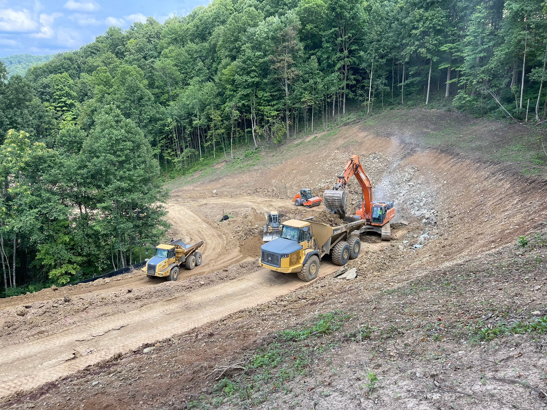 Construction machinery clearing a hillside dirt road beside a forested slope