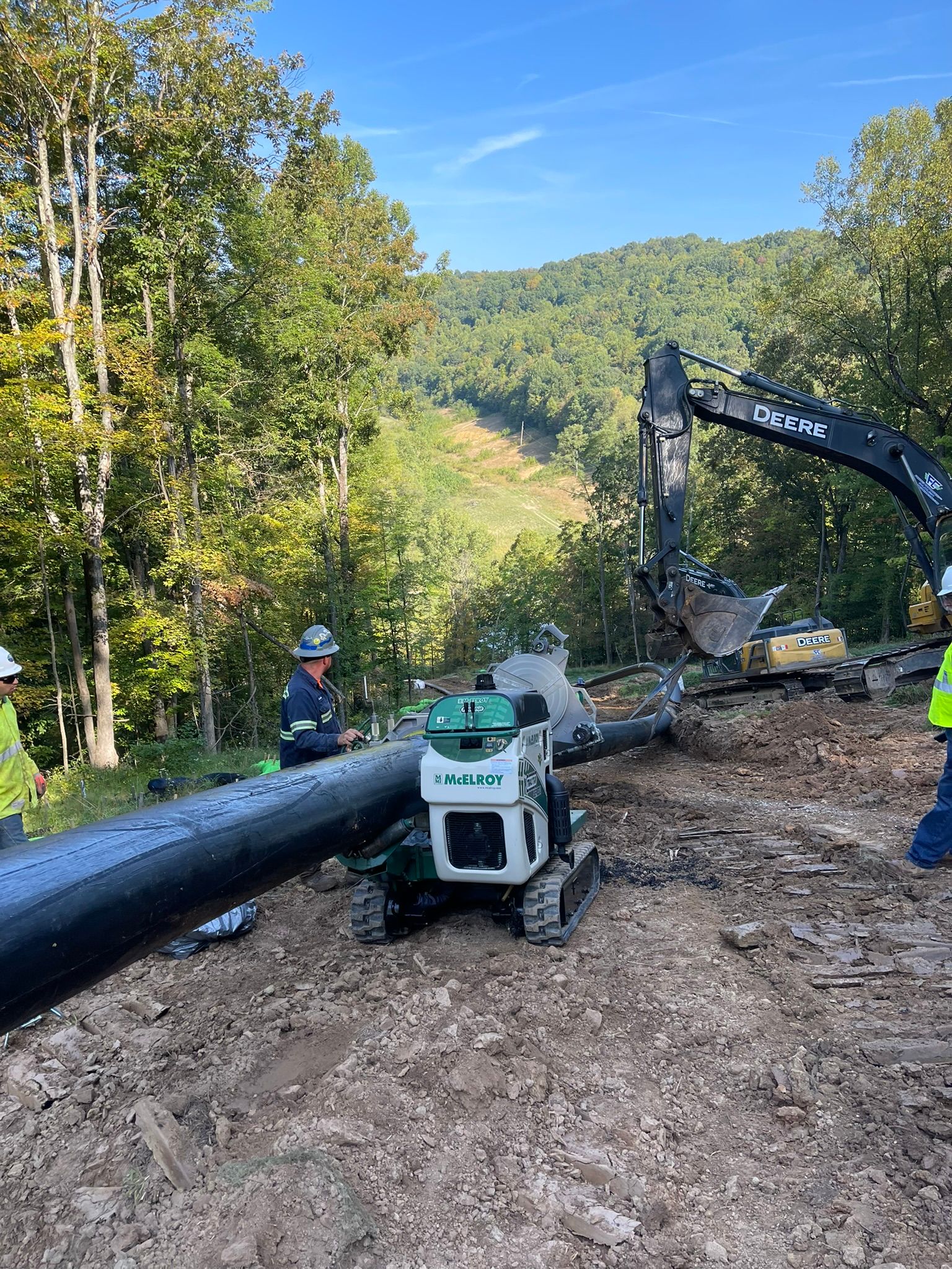 Excavator and skid steer clearing a large pipe on a dirt hillside beside trees and a road
