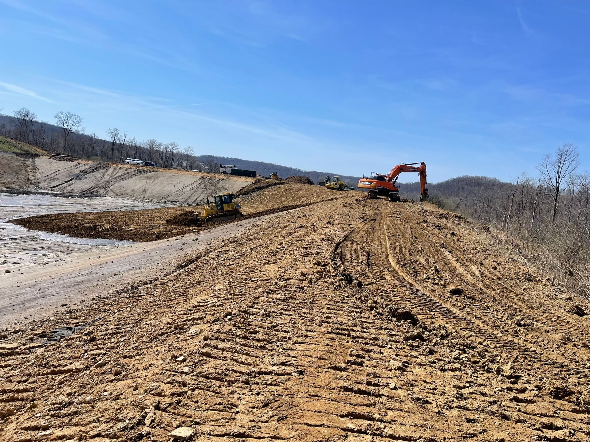 Excavator grading a muddy dirt road on a hillside under a clear blue sky