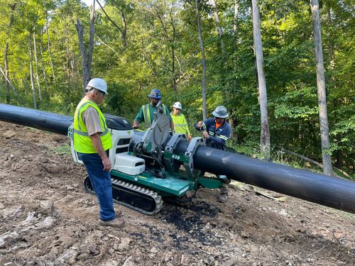 Workers operating a green pipe fusion machine on a black pipe in a wooded construction site