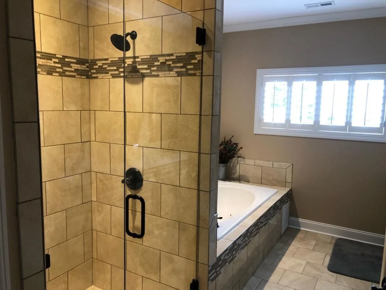 A tiled walk-in shower with a glass door next to a built-in soaking tub in a neutral-toned bathroom with a window.