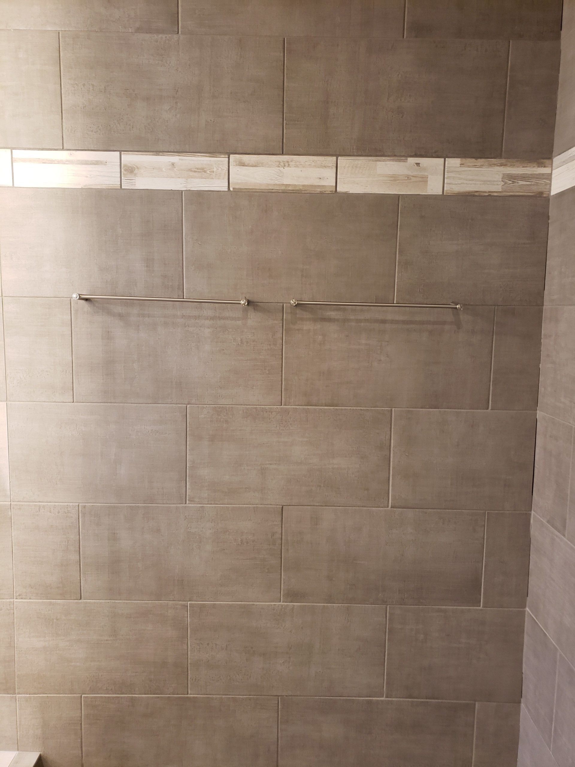 A shower wall with large, rectangular brown tiles, featuring a horizontal row of lighter accent tiles and a metal shelf.