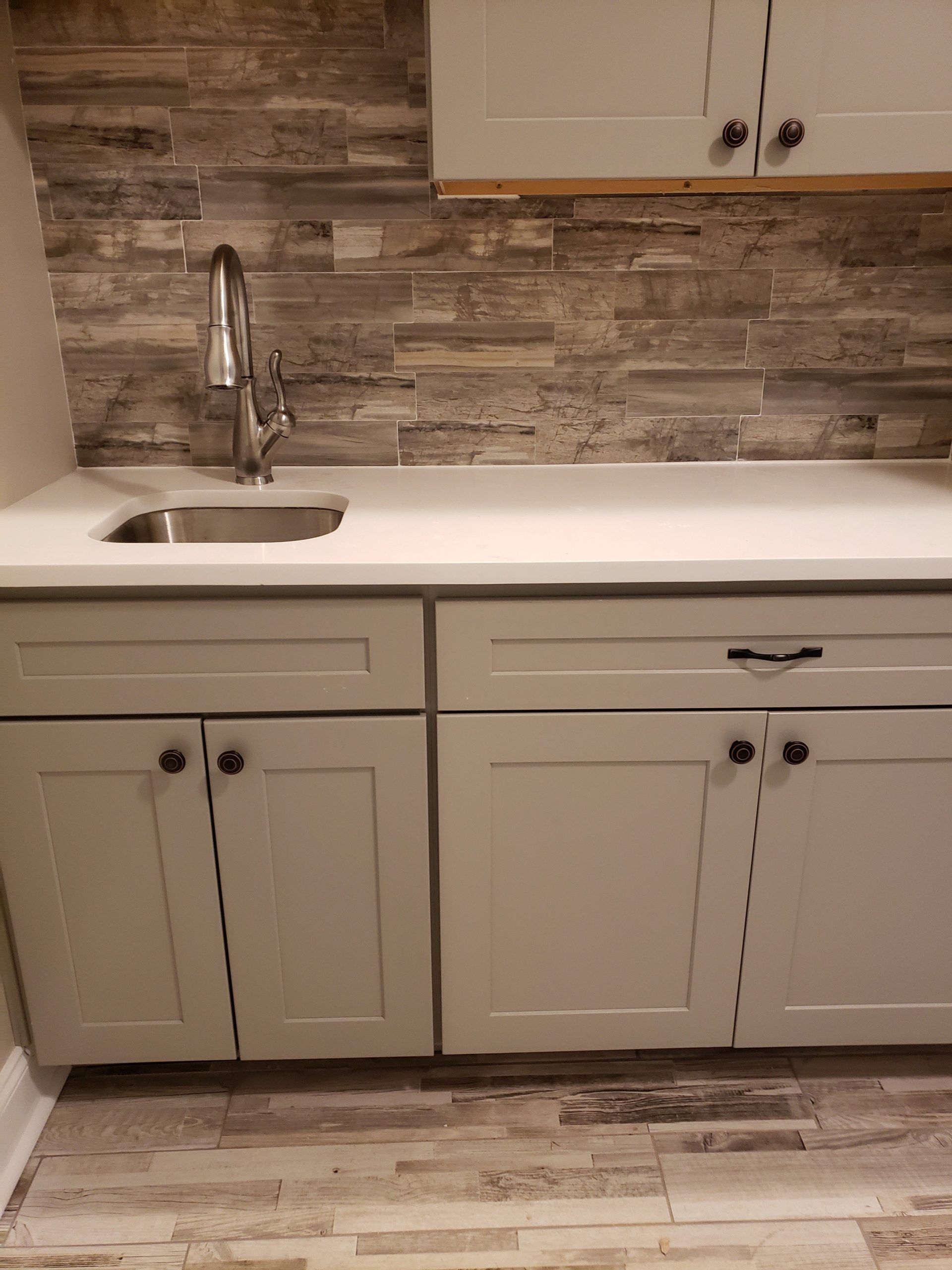 A sink with a chrome faucet set into a white countertop, featuring light gray cabinets and stone-look backsplash and floor.