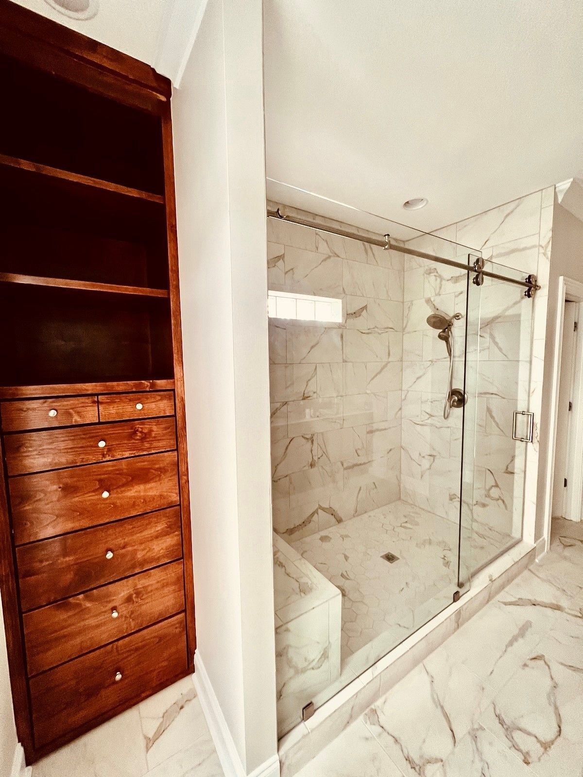 A walk-in shower with marble-style tiles and a glass sliding door, next to a tall wooden shelving unit with drawers.