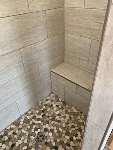 A shower stall interior with light beige rectangular tile walls, a built-in tiled bench, and a multi-colored pebble floor.