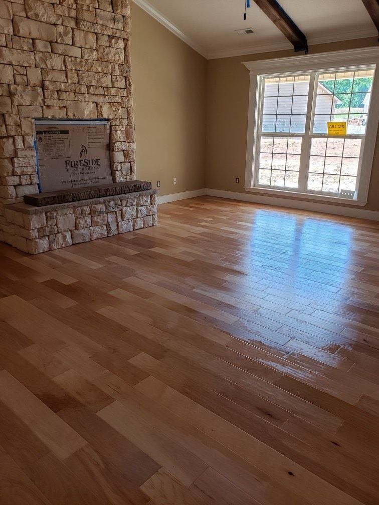 A sunny living room with hardwood floors, tan walls, and a large stone fireplace with a wooden mantel.