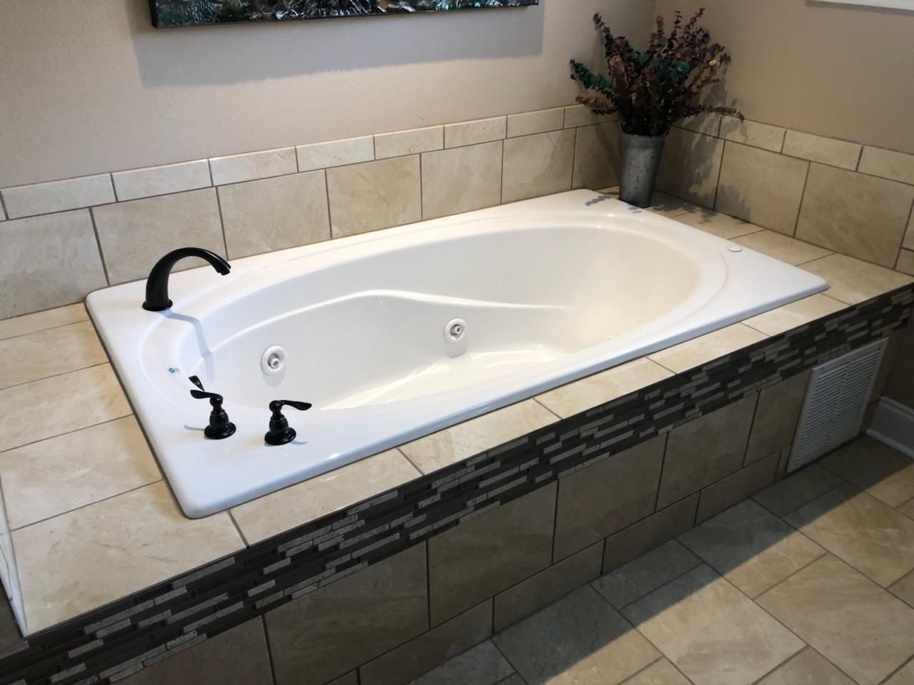 A white built-in soaking tub with black fixtures, surrounded by beige and dark stone tiles, in a bathroom.