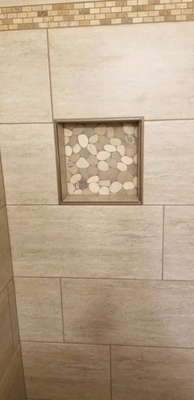 A square shower niche with pebble tile inlay, set against large, beige, textured horizontal wall tiles.