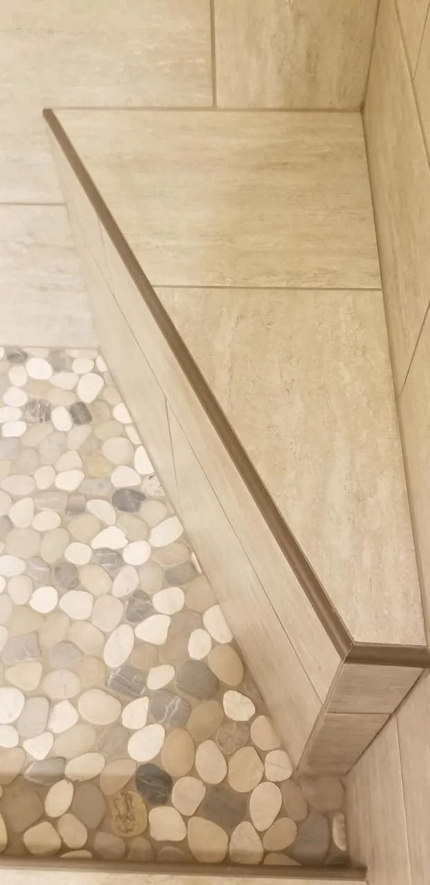 A corner shower bench tiled in beige stone, accented with a dark trim, situated above a pebble-stone floor.
