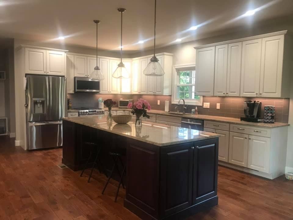 A kitchen featuring a dark island with seating, white upper cabinets, stainless steel appliances, and wood flooring.