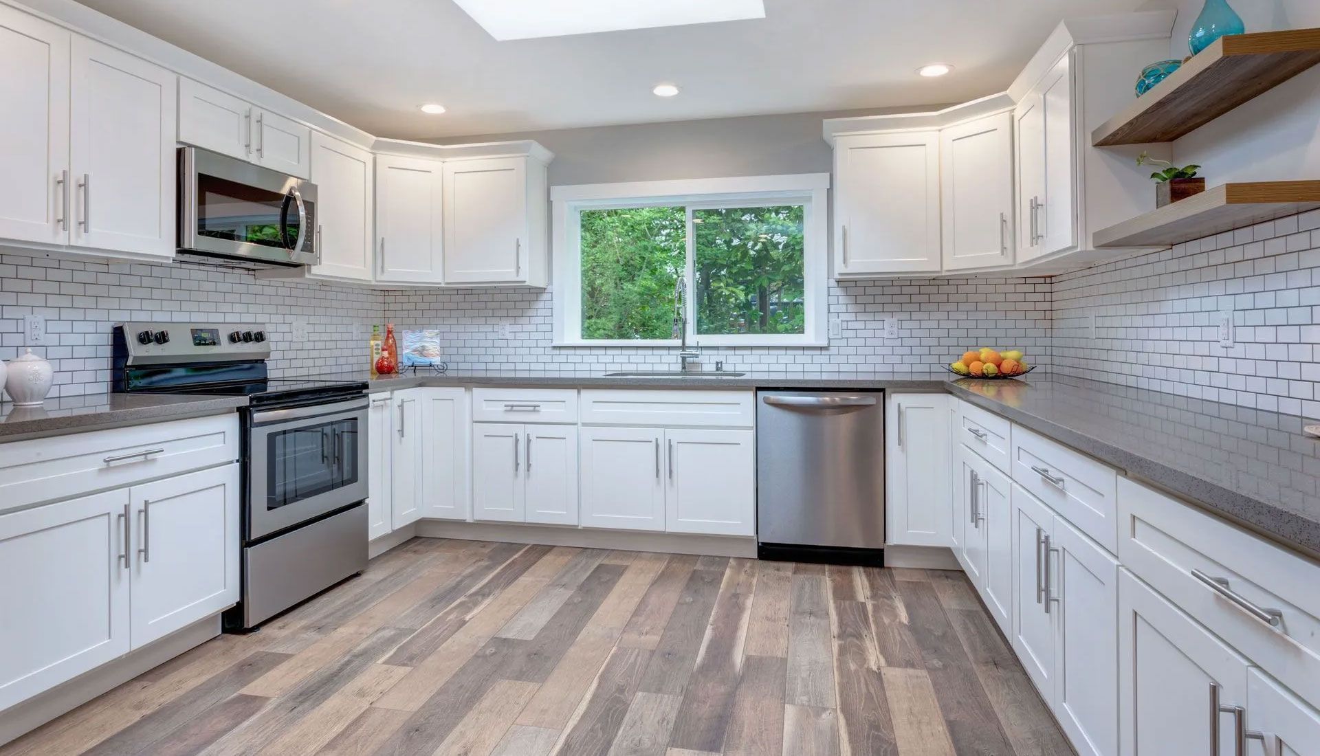 A white kitchen with shaker cabinets, stainless steel appliances, dark countertops, a tile backsplash, and wood floors.