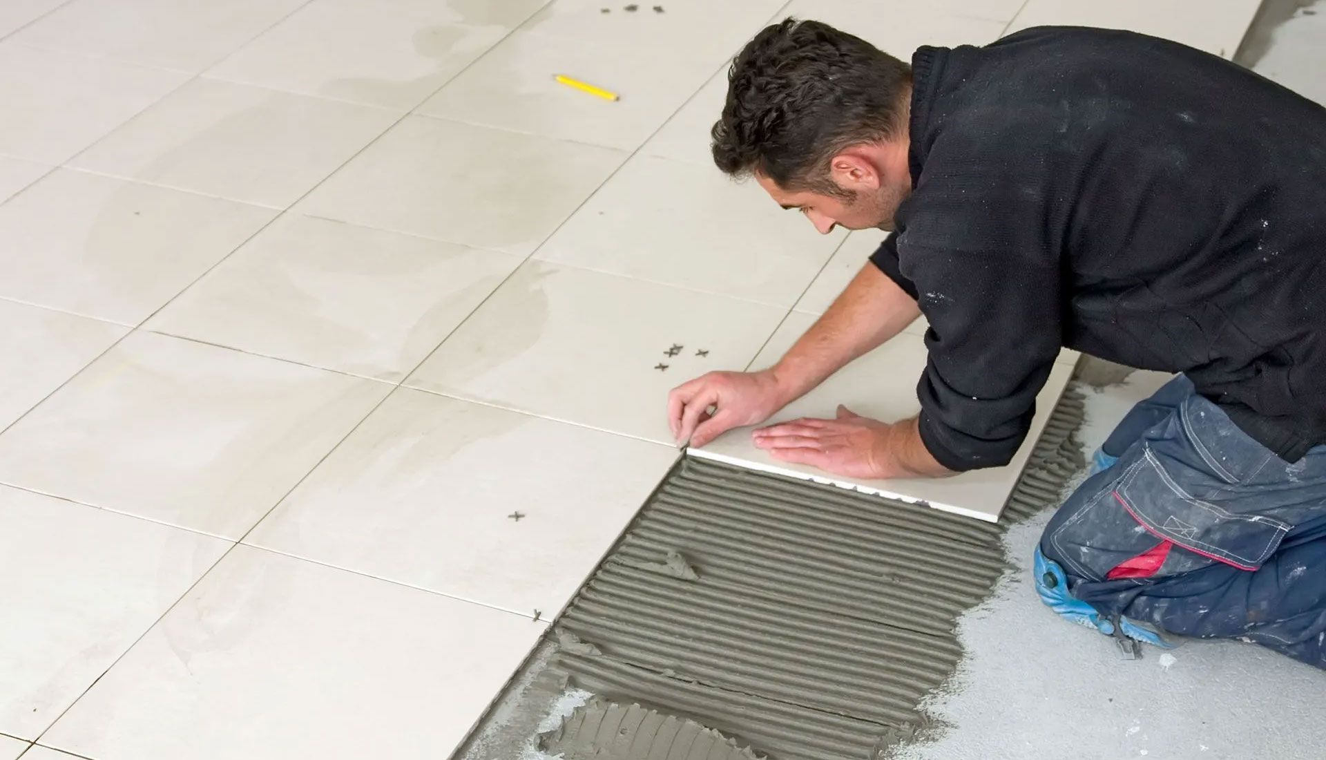 A person kneeling on a floor, carefully laying a large light-colored tile onto a bed of ridged adhesive.