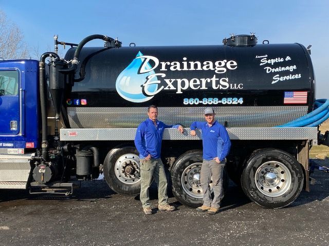 Two men in blue jackets stand in front of a black and silver Drainage Experts truck.