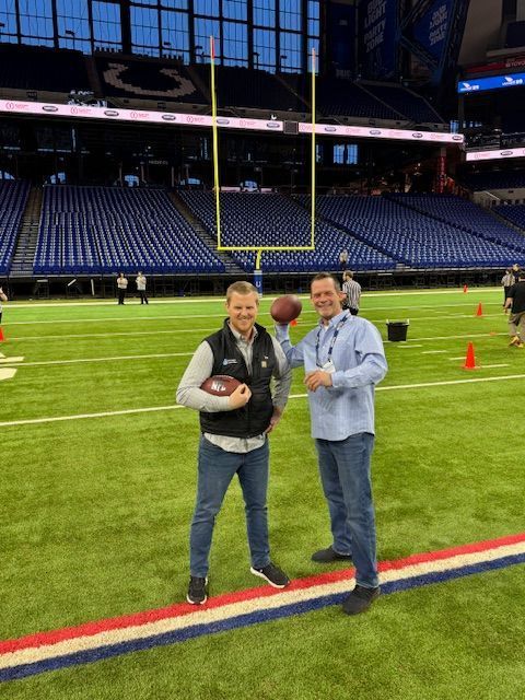 Two men on a football field, holding footballs near a goalpost, inside a stadium.